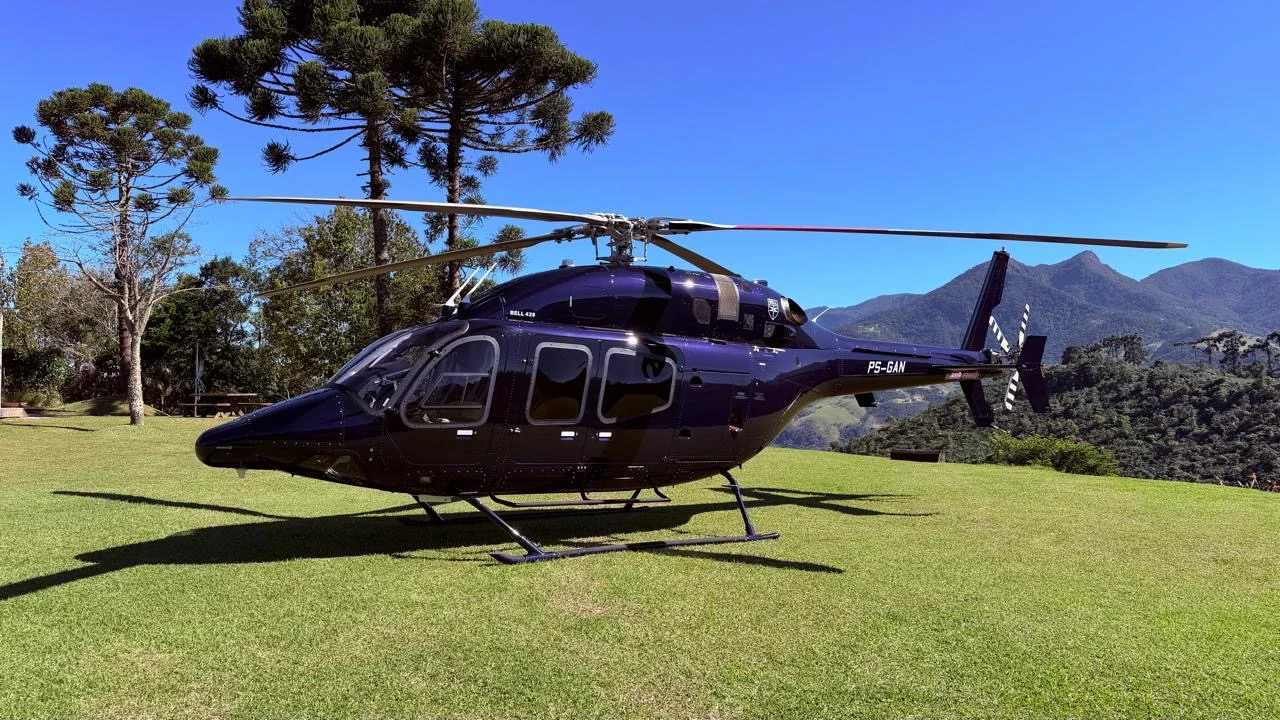 Bell 429 azul escuro pousado na grama entre araucárias e montanhas